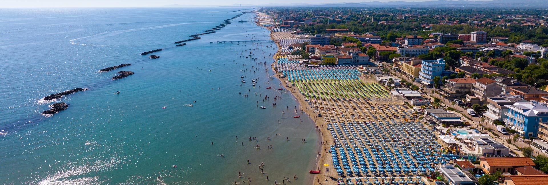 Veduta aerea della lunga spiaggia di San Mauro Mare: file ordinate di ombrelloni colorati si estendono lungo la costa, con molte persone in acqua e sul bagnasciuga; il mare calmo è protetto da scogliere frangiflutti e la città si sviluppa alle spalle dell